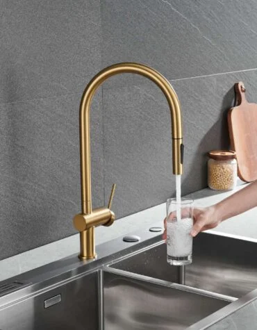 Torneira de cozinha elegante Berna com acabamento ouro mate, bica alta curvada e manípulo lateral, instalada numa bancada dupla em inox com fundo de pedra escura, enquanto um copo é enchido com água.